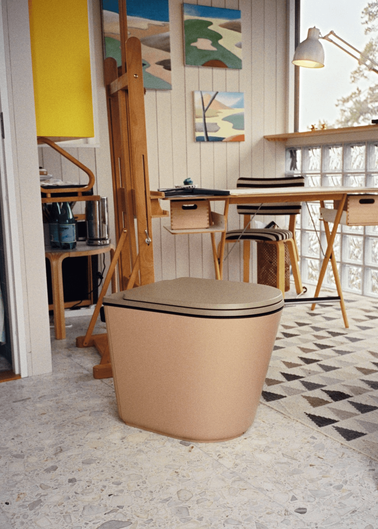 Beige komposttoalett i ljust konstnärsrum med staffli, tavlor och glasblocksvägg