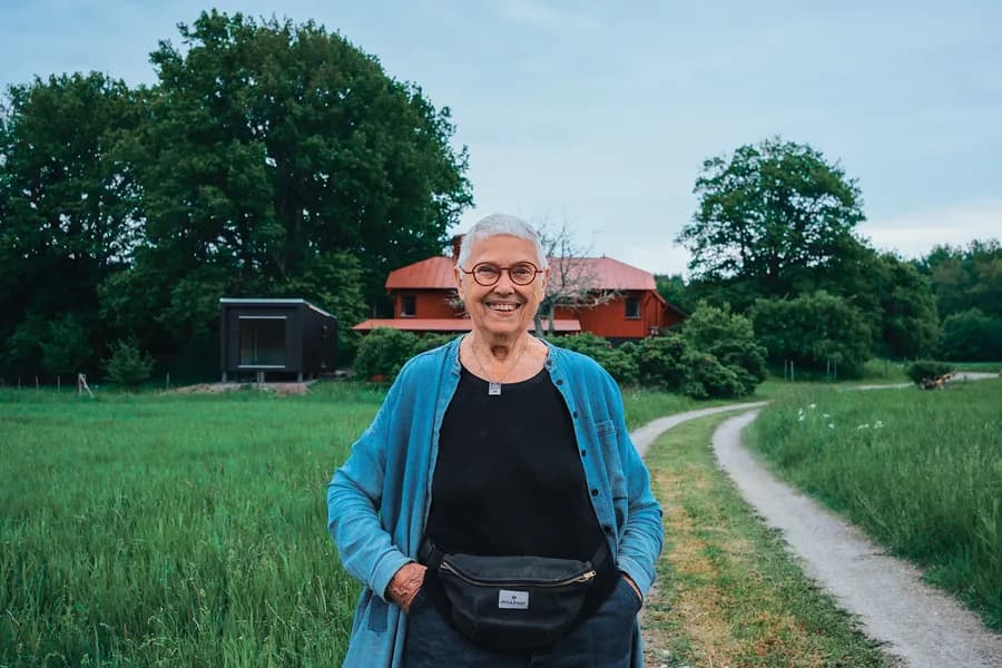 Older woman with red glasses on a path with a red cottage and green field behind her