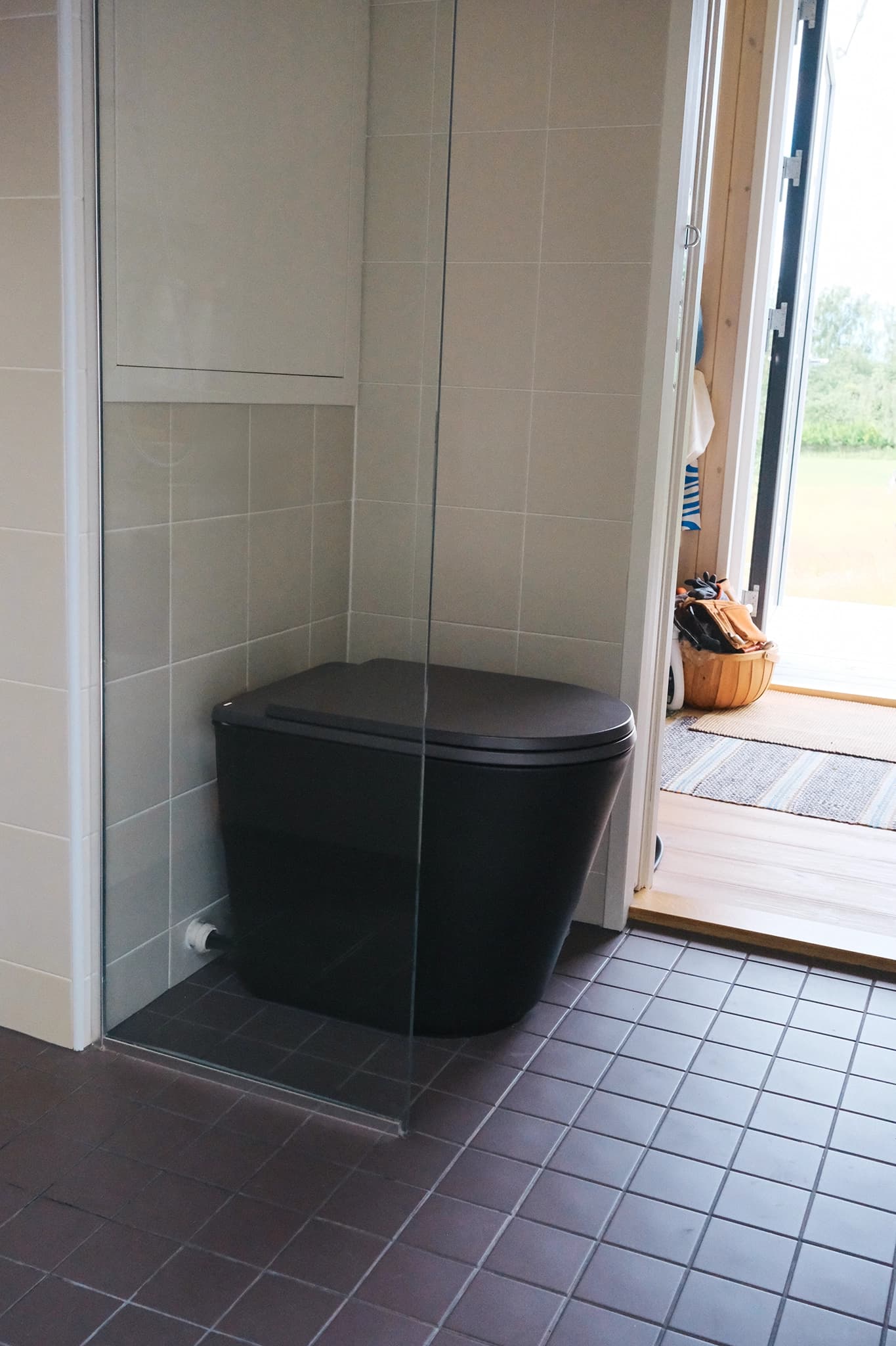 Black Luna dry toilet behind a glass partition on dark tile flooring in a light tiled bathroom