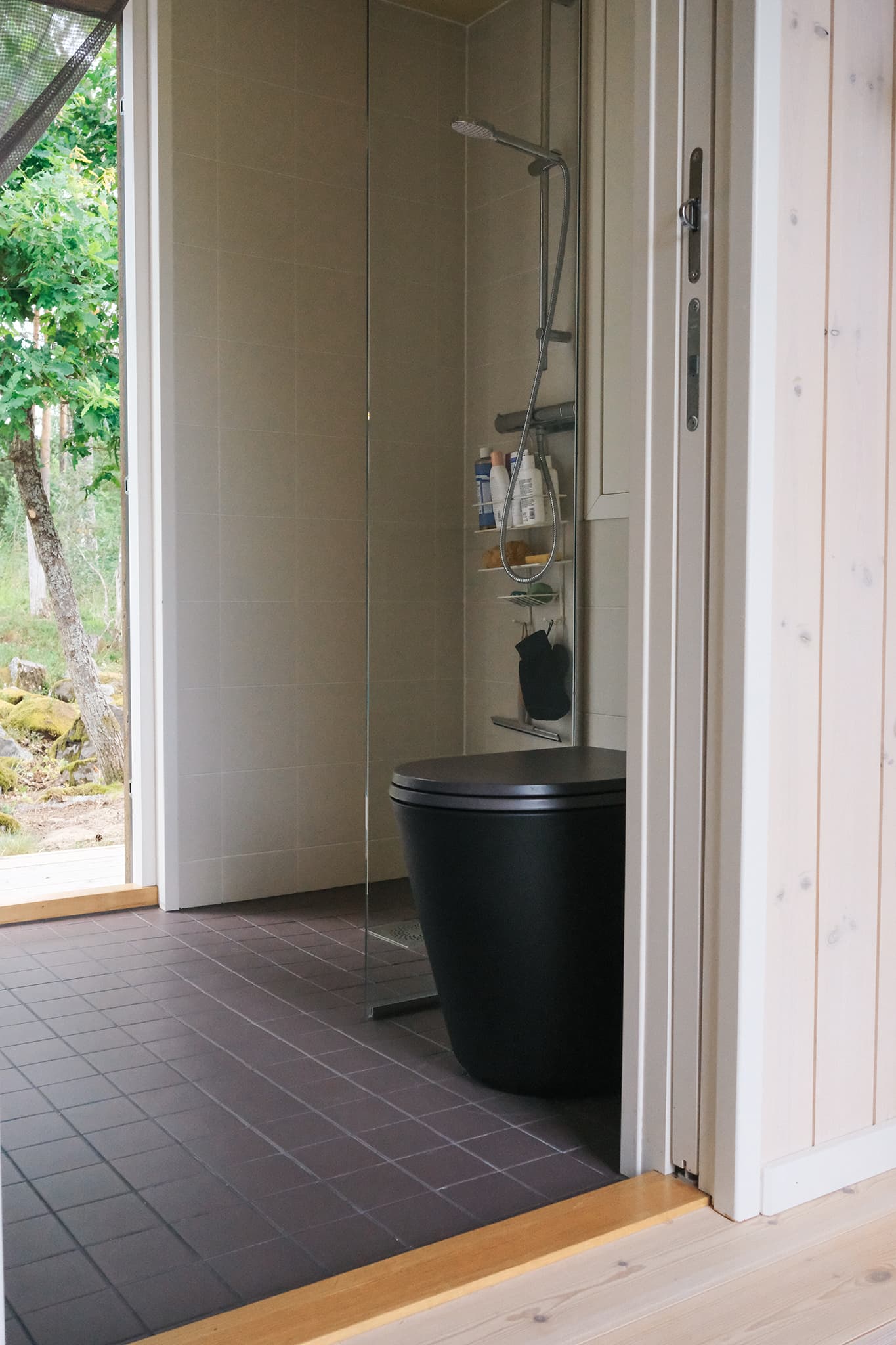 Black Luna compost toilet in a bright bathroom with shower, dark floor tiles and forest view