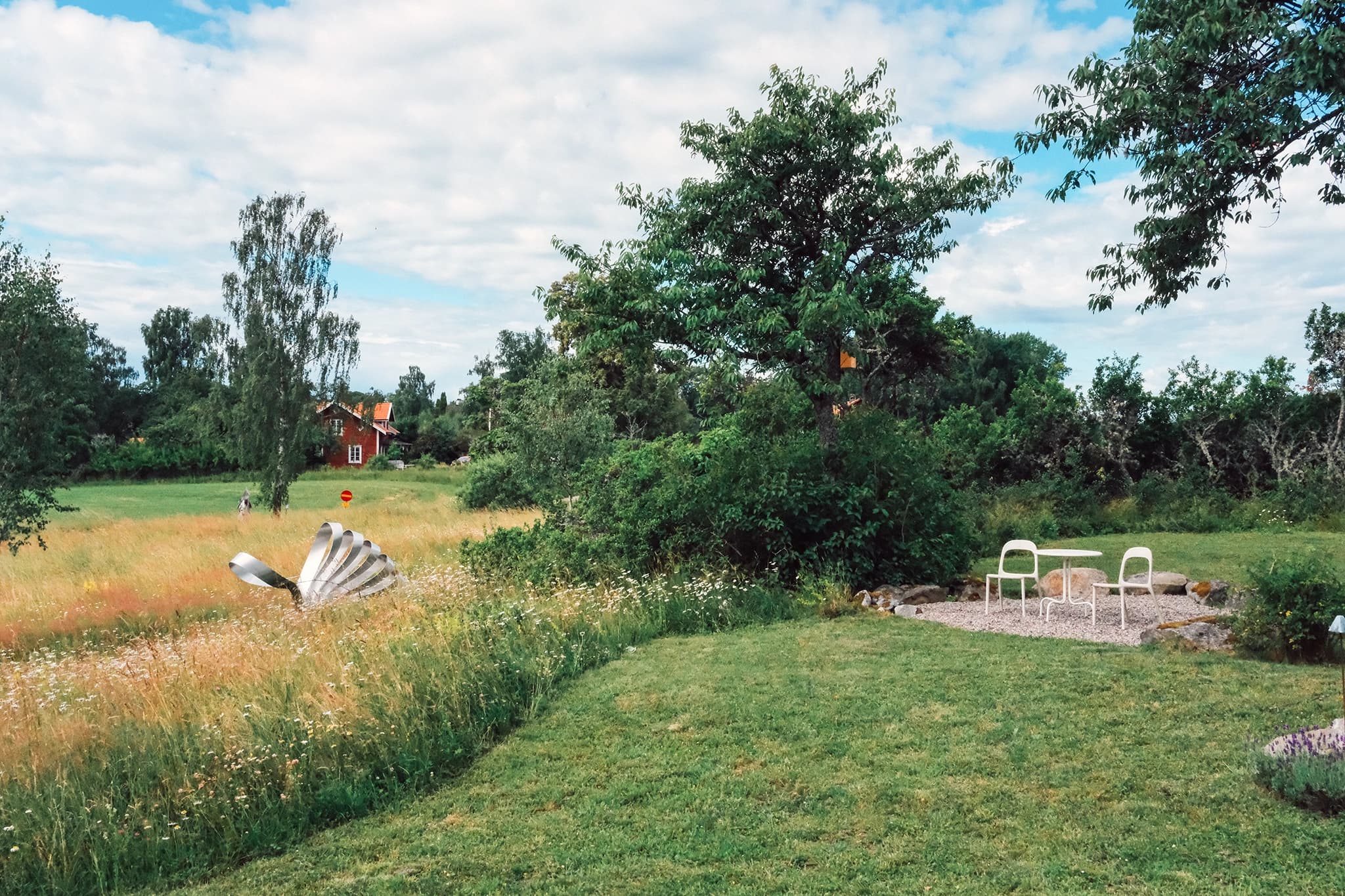 Green garden with lawn, wildflower meadow, white table and chairs, and a red cottage in the distance
