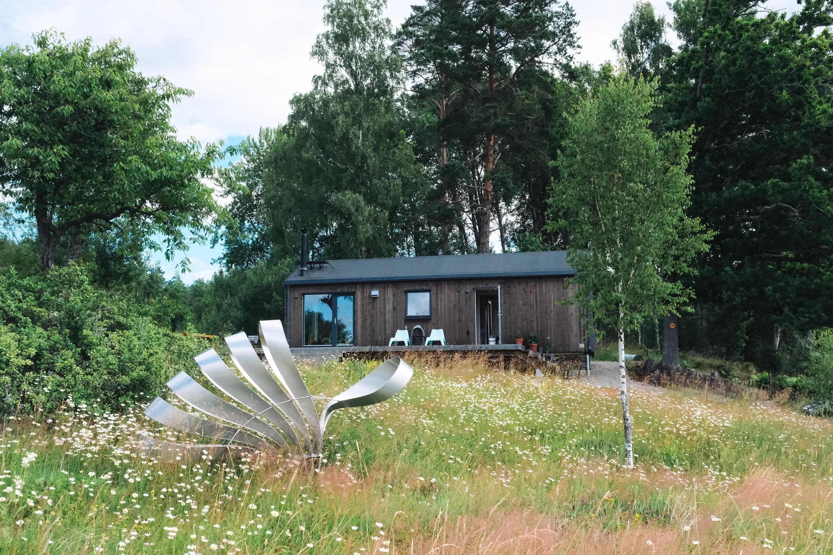 Mörk trästuga på grässluttning med metallskulptur och vildblommor i förgrunden